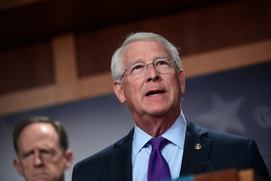 Roger Wicker, dressed in a jacket and tie, stands in front of a wall with a star on it.