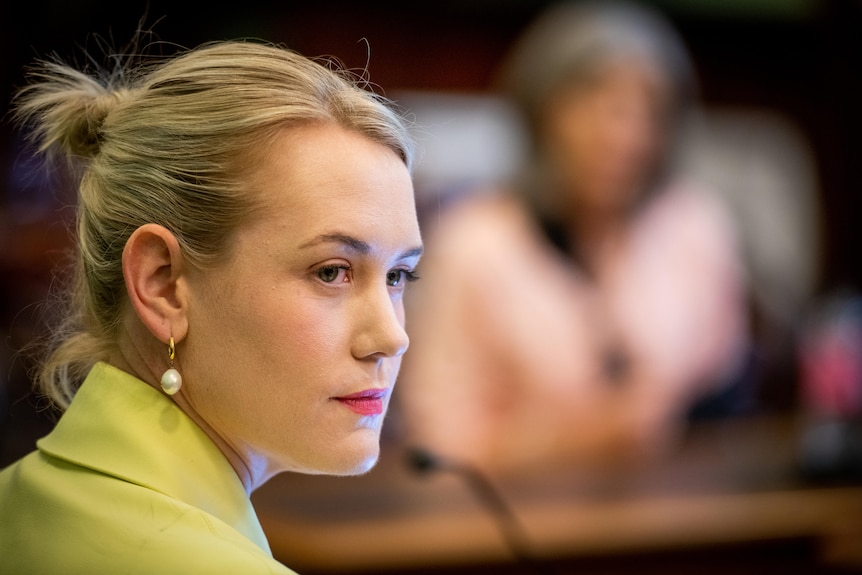 A woman wearing a yellow jacket and pearl earrings sits in front of a small microphone 