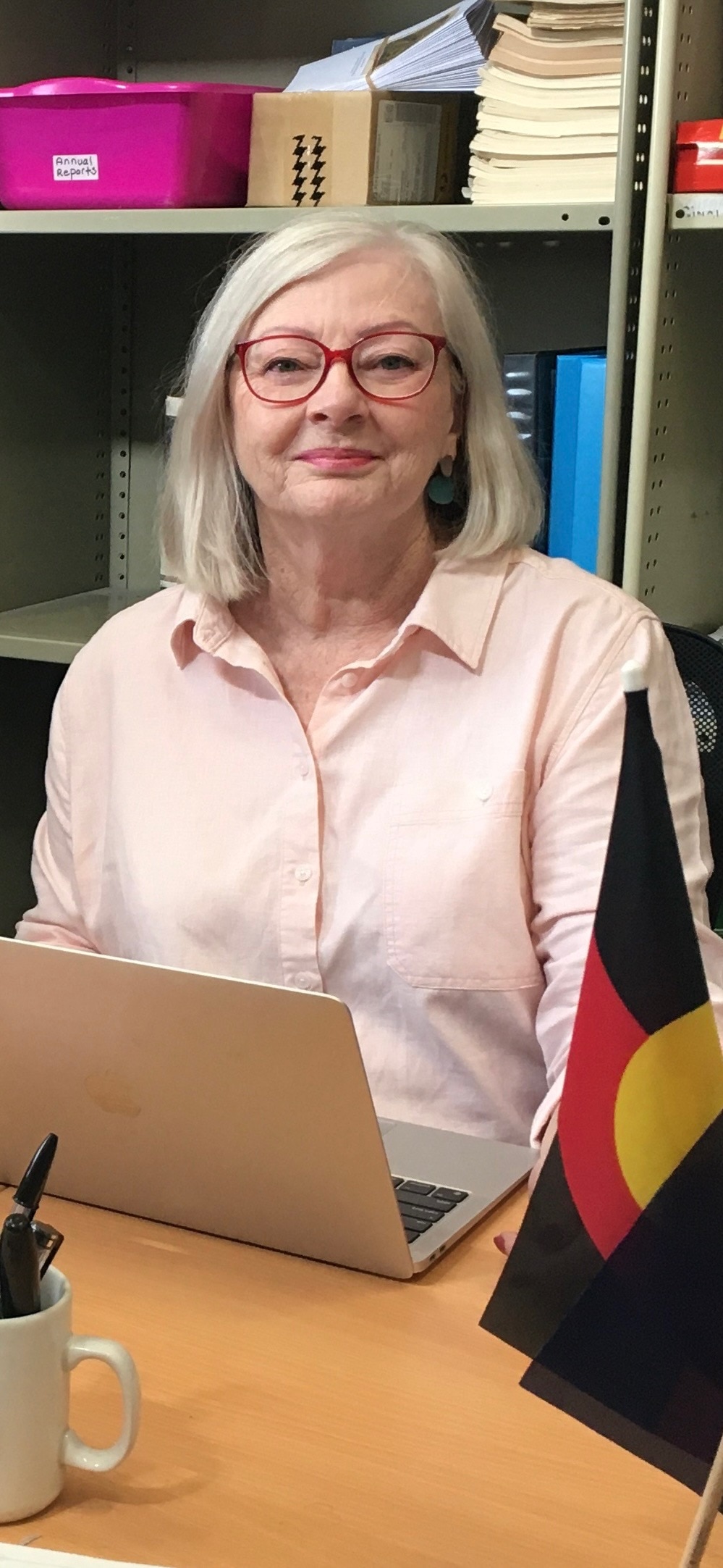A woman with white hair, wearing glasses, sits at a laptop. An Aboriginal flag sits in front of her.