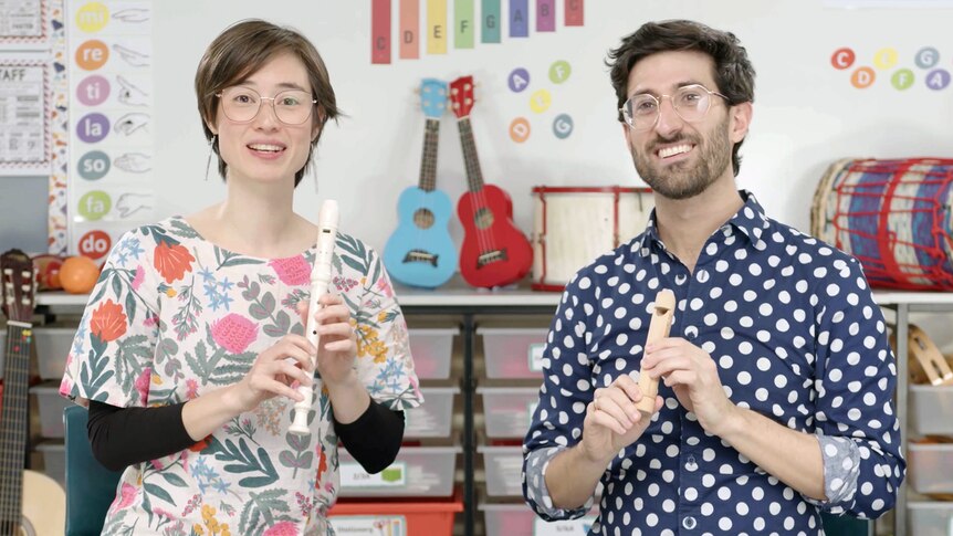 Two smiling primary school teachers sit in a decorated music classroom holding a recorder and a slide whistle.