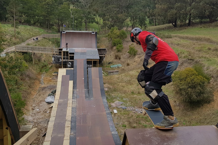 A skateboarder wearing a red t-shirt about to roll down a giant skate ramp built in a gully.