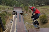 A skateboarder wearing a red t-shirt about to roll down a giant skate ramp built in a gully.