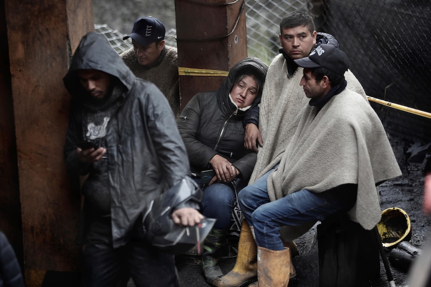 People sitting outside in the rain lean on each other as someone in front looks at their phone. 