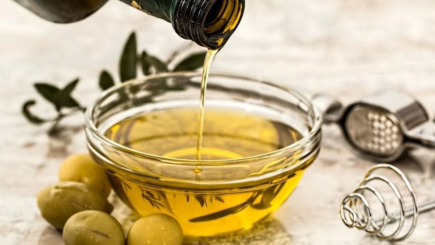 Glass jar filled with olive oil with green olives next to it.