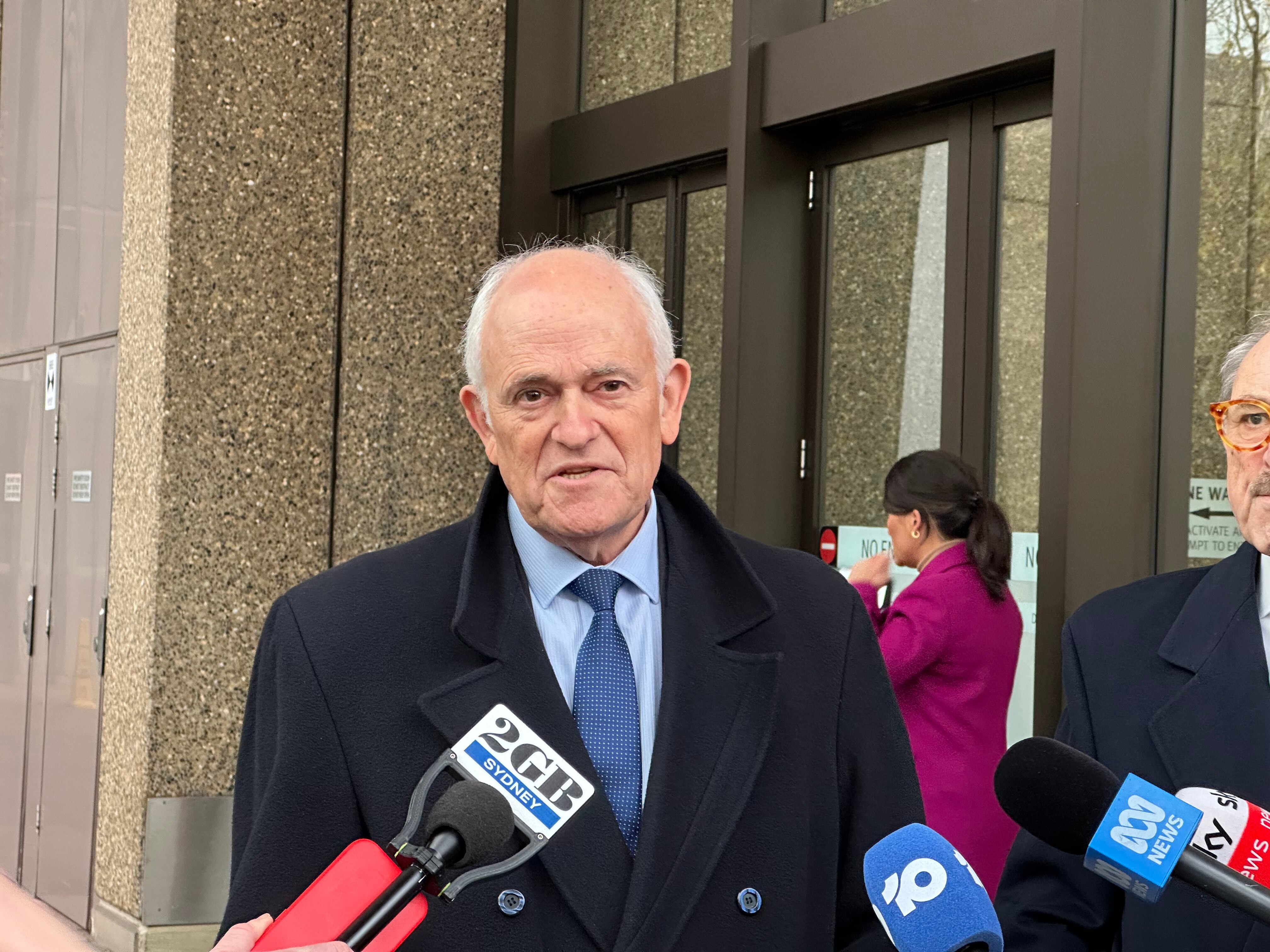 An older man wearing a blue tie and light blue shirt gives grabs outside a courthouse