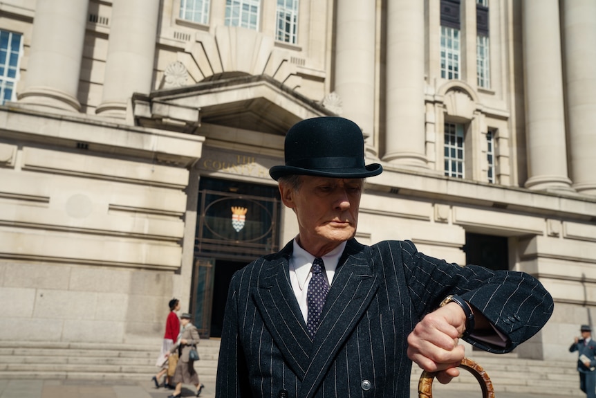 A 70-something man in a 50s-style bowler hat and pinstripe suit looks at his watch as he stands outside a town hall