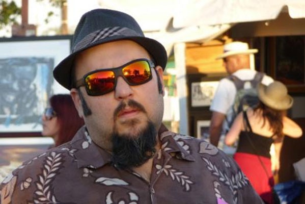 Photo of AJ Maddah, he is wearing a fedora and wraparound sunglasses.