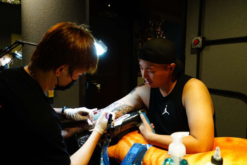 A tattoo artist wears a mask as he gives a tattoo to a man wearing a singlet.