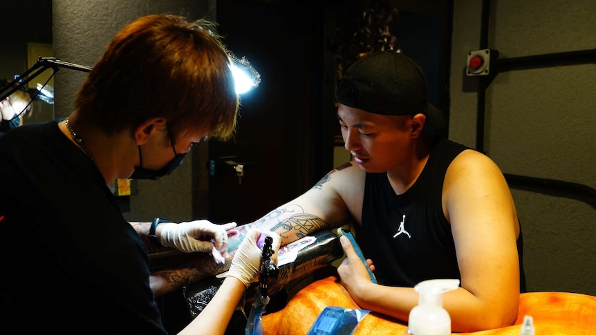 A tattoo artist wears a mask as he gives a tattoo to a man wearing a singlet.