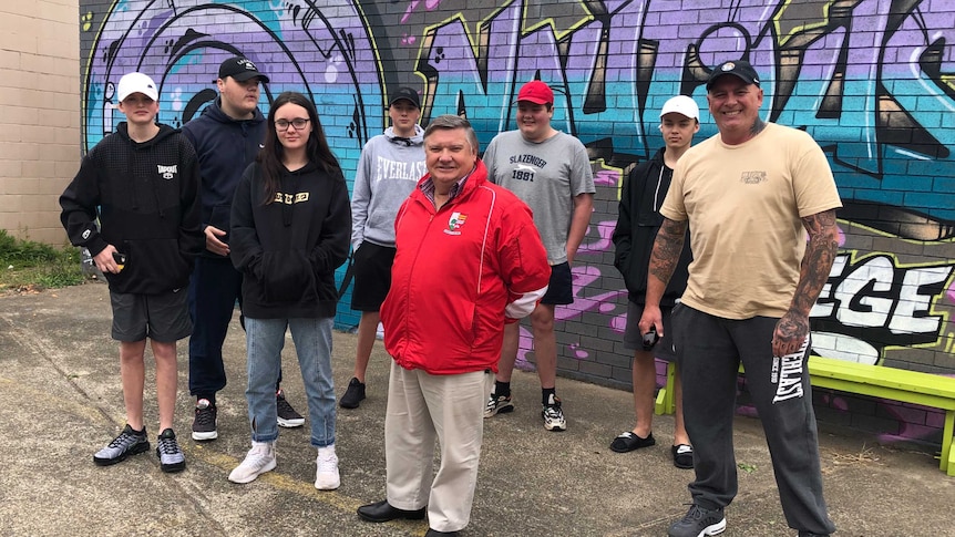 A group of teenagers and two male teachers stand inside a wall decorated in purple and blue graffiti