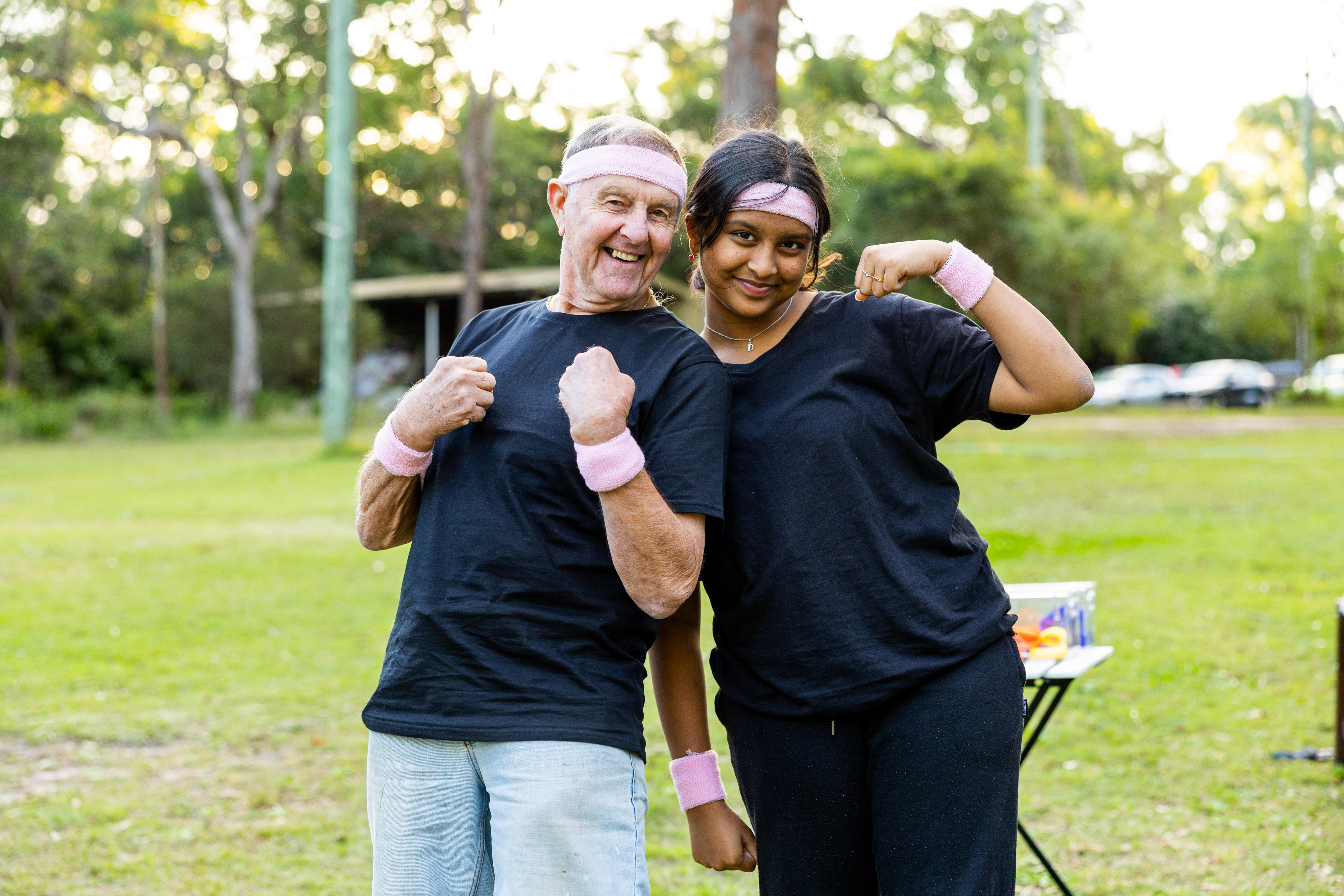 Senior person, Dave, and teen, Vya, wearing pink head and sweat bands flex their muscles in an outdoor park