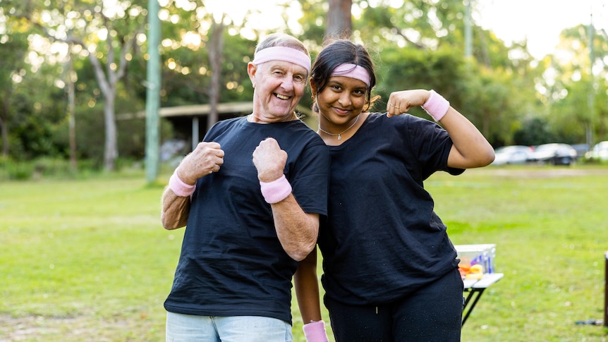 Senior person, Dave, and teen, Vya, wearing pink head and sweat bands flex their muscles in an outdoor park