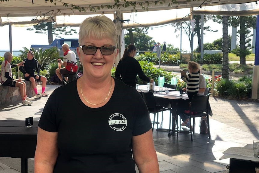 A woman wearing sunglasses smiles as she poses for a photograph in an outside dining area where people sit in the sunshine