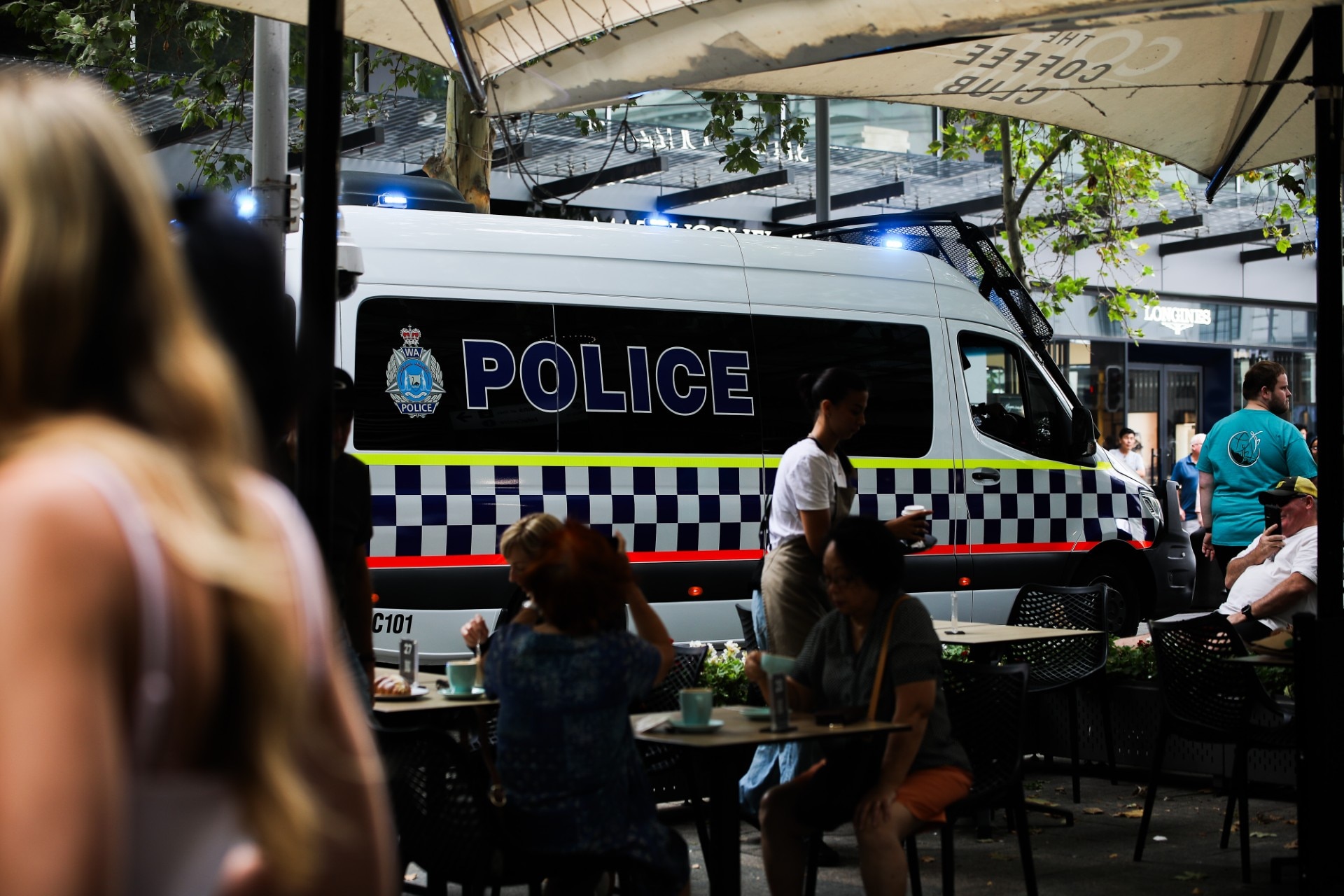 Un coche de policía al otro lado de gente sentada en mesas al aire libre.