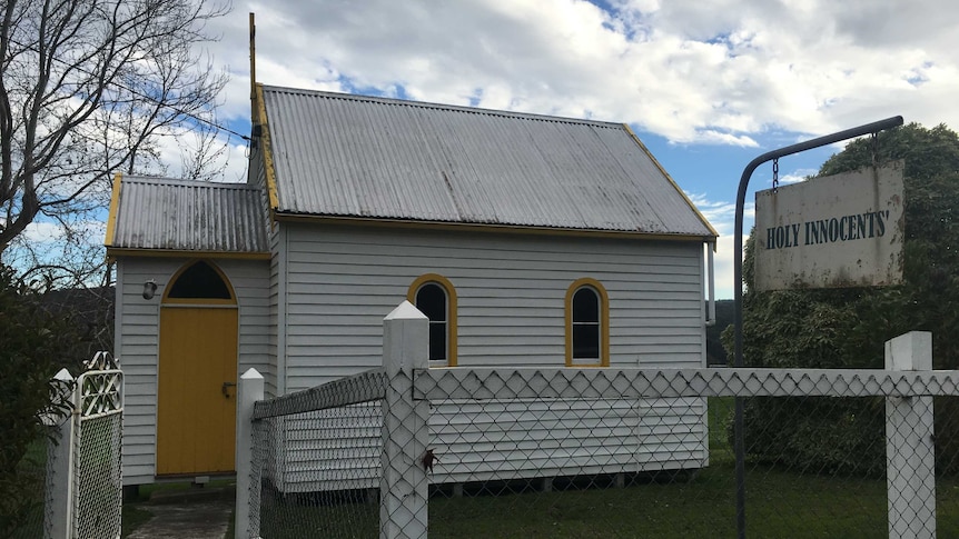 A tiny timber church, made from timber, with a tin rood, pictured from the exterior. A sign out front reads 'Holy Innocents'.