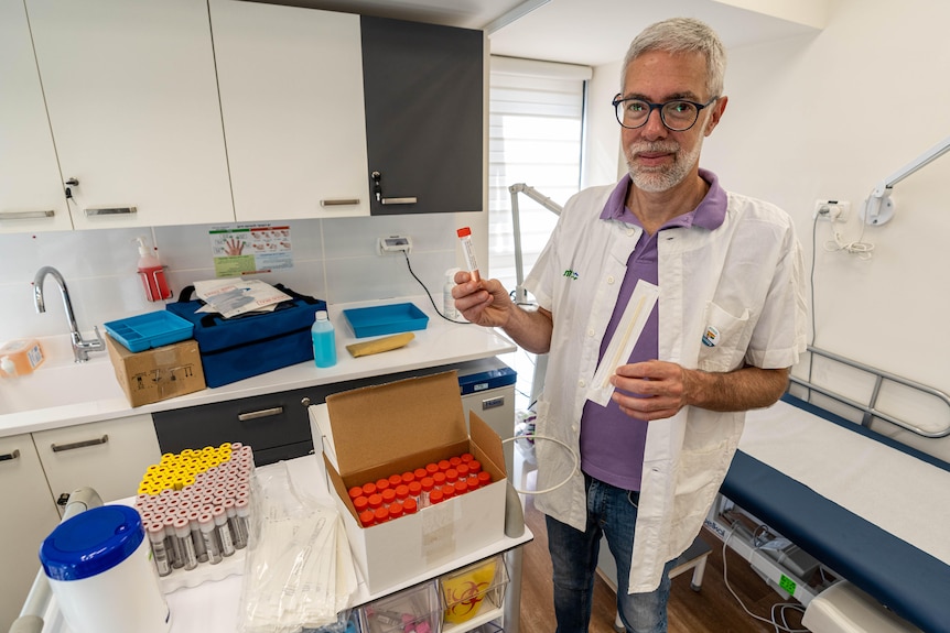 A man with a gray beard dressed in a lab coat stands in a GP's office holding up a vial from a big box of vials 