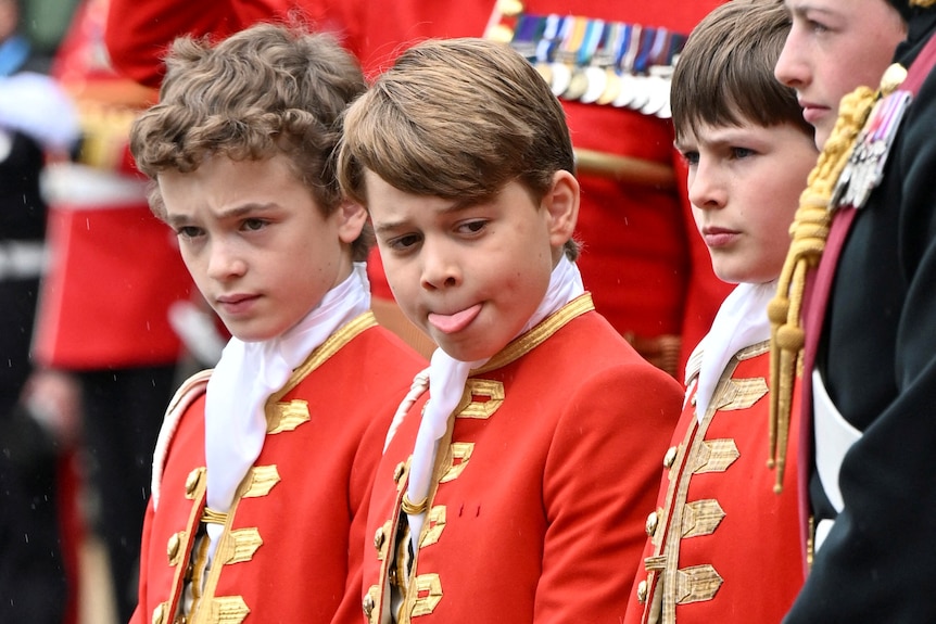 Three boys in formal red jackets standing, George is in the centre with his tongue out