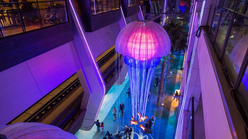 Giant jellyfish with pink bodies and blue tentacles made of lights hang from the ceiling with people walking below.
