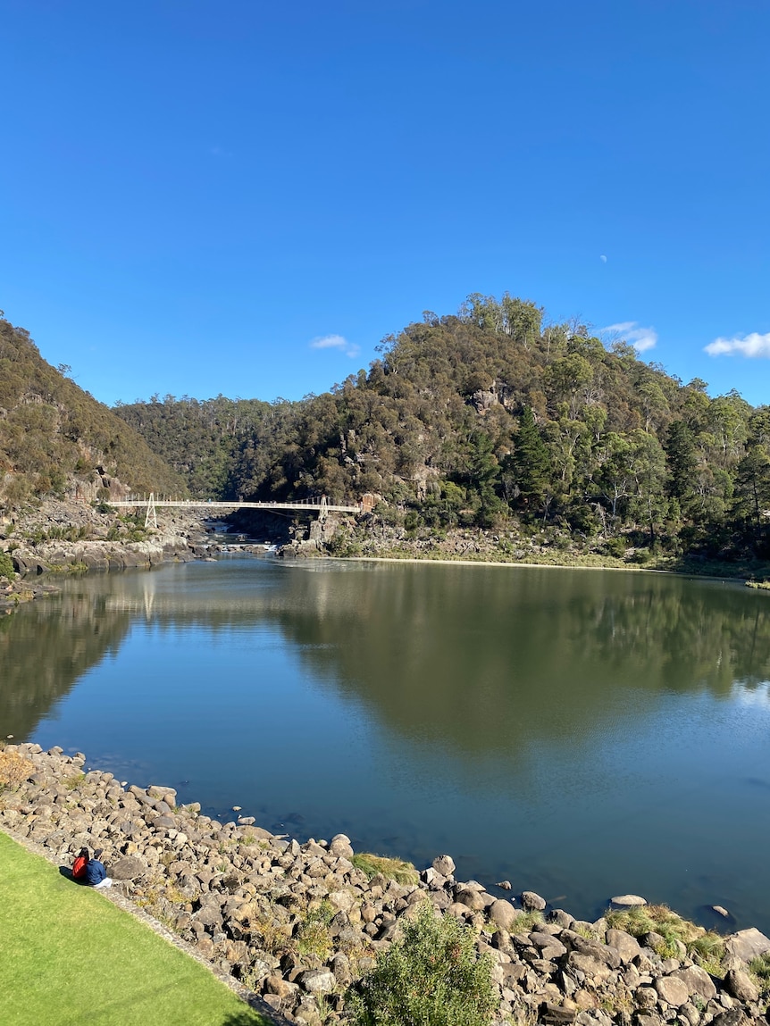 A landscape in Launceston with a bridge and pool of blue water over a blue sky. Deeply forested mountains  in the background.