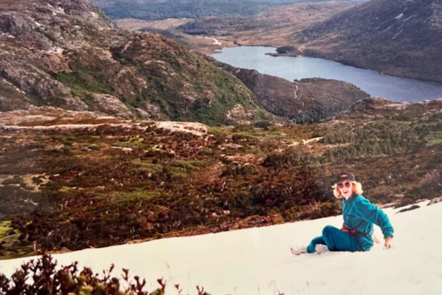 Old photo of blonde-haired woman wearing blue tracksuit sitting on the side of a mountain, beaming.