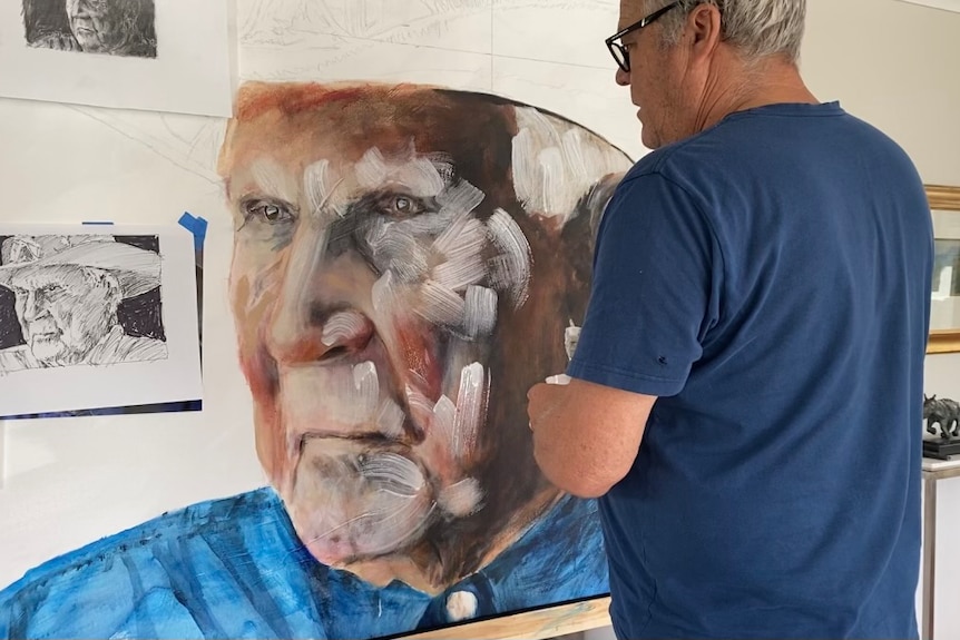 An artist paints a large portrait of an Indigenous man wearing a blue collared shirt.