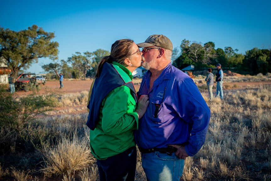 Karen and Angus Emmott share a kiss on their property, with dry grass and green trees surrounding them.