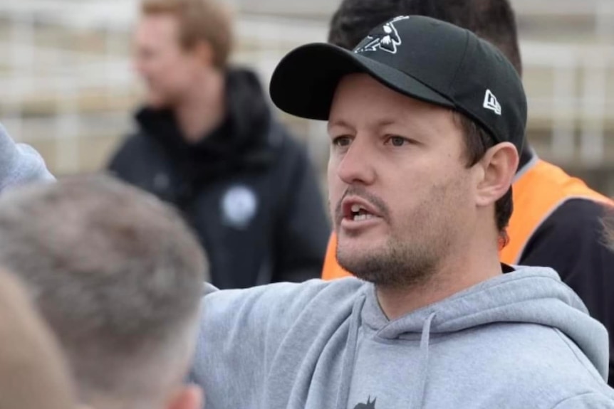 A man wearing a black hat and a grey hoodie pointing 