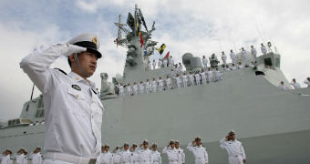 Sailor stands saluting in the foreground and group of saluting sailors stand before and on deck of a naval boat.