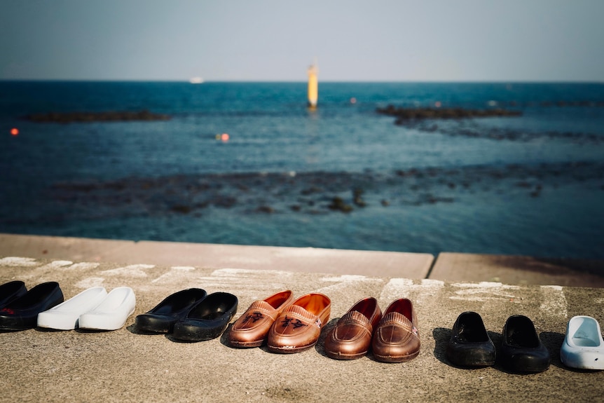 A group of shoes in different colours lined up on cement beside the sea.
