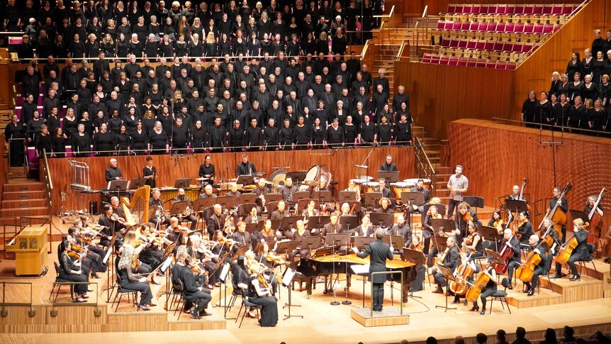 The stage and seating behind is full of singers and orchestral players in the brightly lit concert hall at Sydney Opera House.