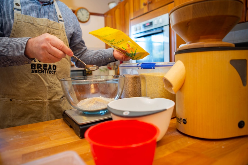 A man's hand measures seeds into a mixing bowl in a busy home kitchen