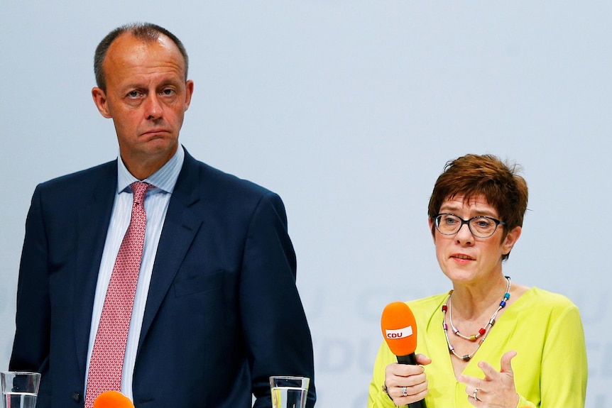 Two tall men and one quite short women stand on stage, the woman is holding an orange microphone.