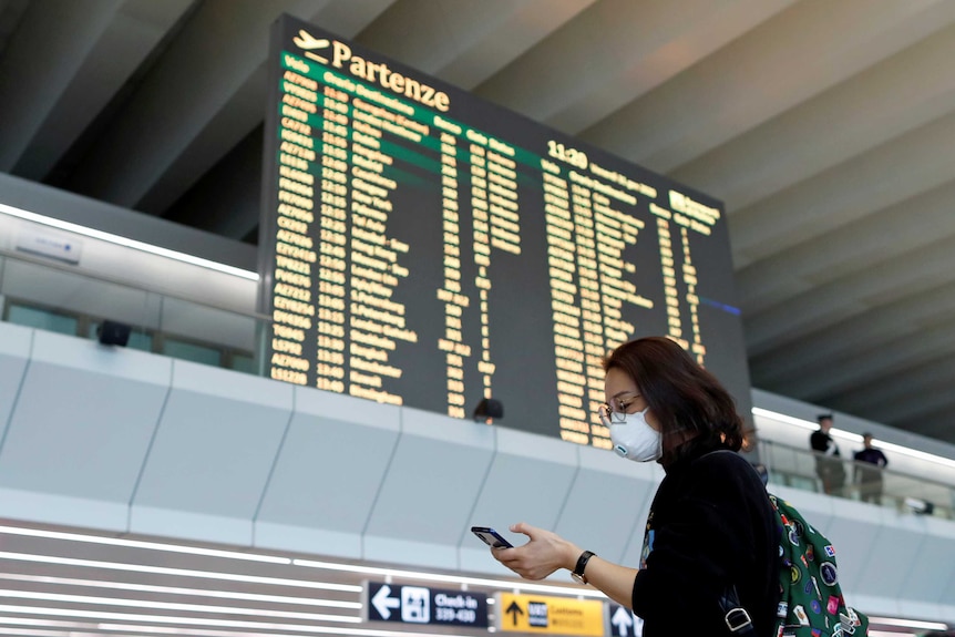 A woman in a face mask checks her phone in a deserted airport with a departure screen behind her