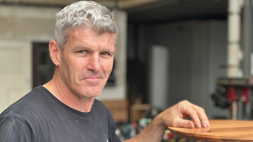 Former AFL player Shaun Smith stands with his hand on a bedside table in a workshop.