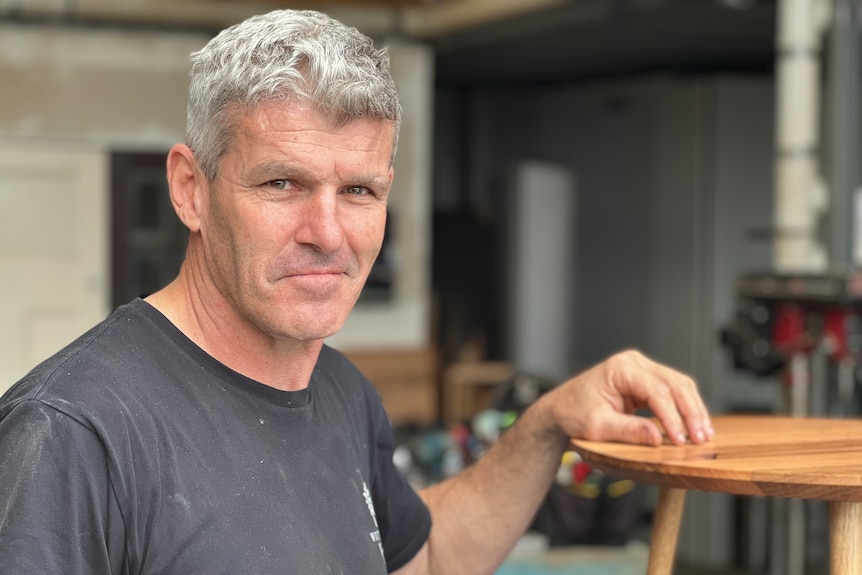 Former AFL player Shaun Smith stands with his hand on a bedside table in a workshop.