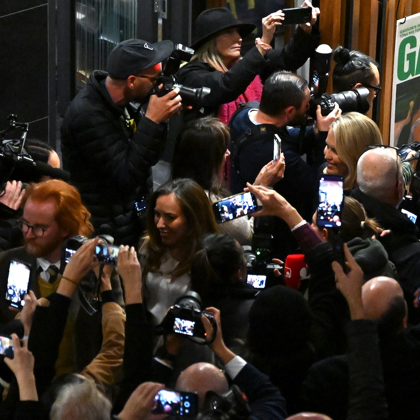 A high shot of a crowd of people, many holding phones to photograph a woman walking through them.