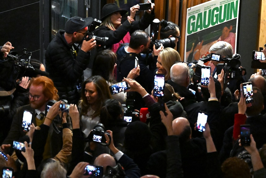 A high shot of a crowd of people, many holding phones to photograph a woman walking through them.