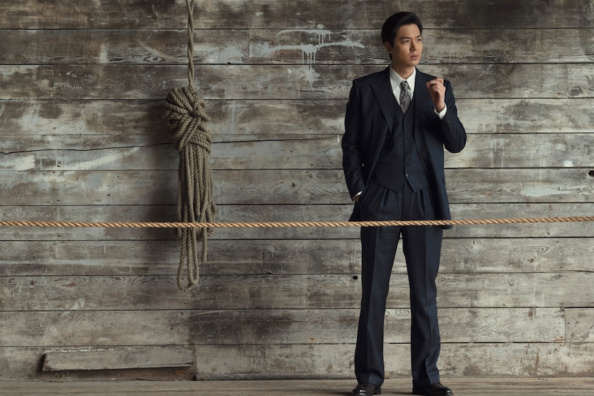 A young Korean man stands in a sharp black suit stands in front of a wooden wall. 