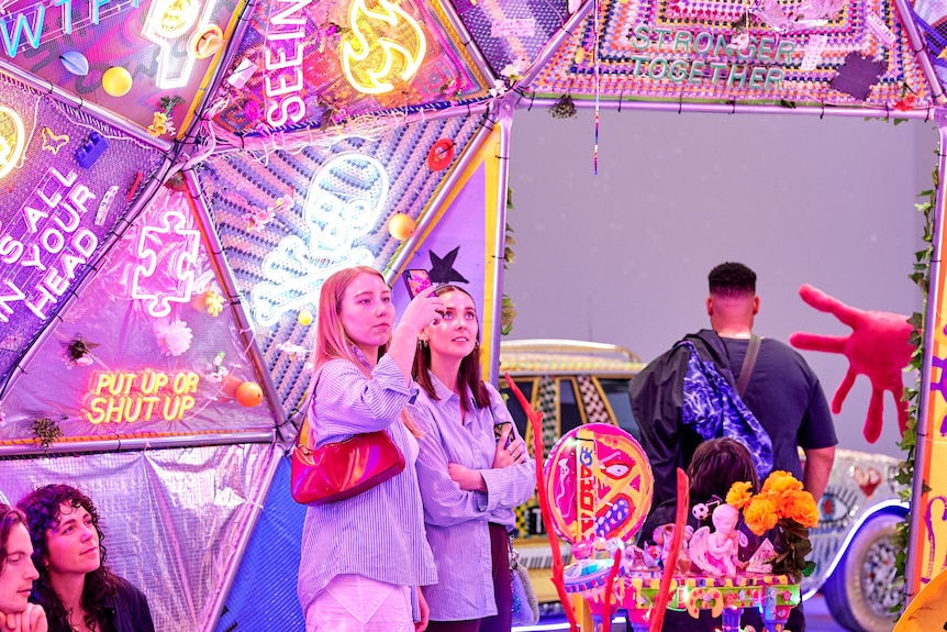 Two people stand in a neon-lit artistic installation, one holding a mobile phone.