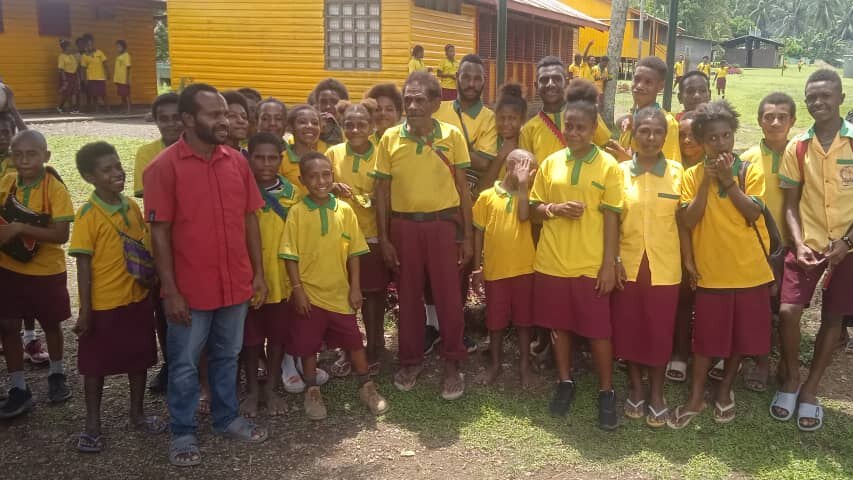 A group of young children and two men, one dressed in the school colours of yellow and red, stand together smiling.