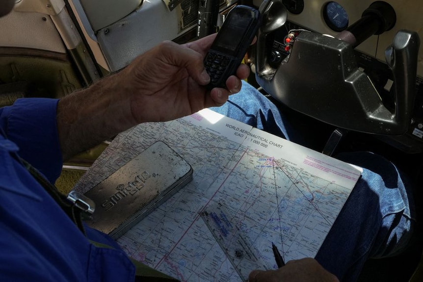 A man holds a phone out over a map, which rests on his lap in the cockpit of a small plane 