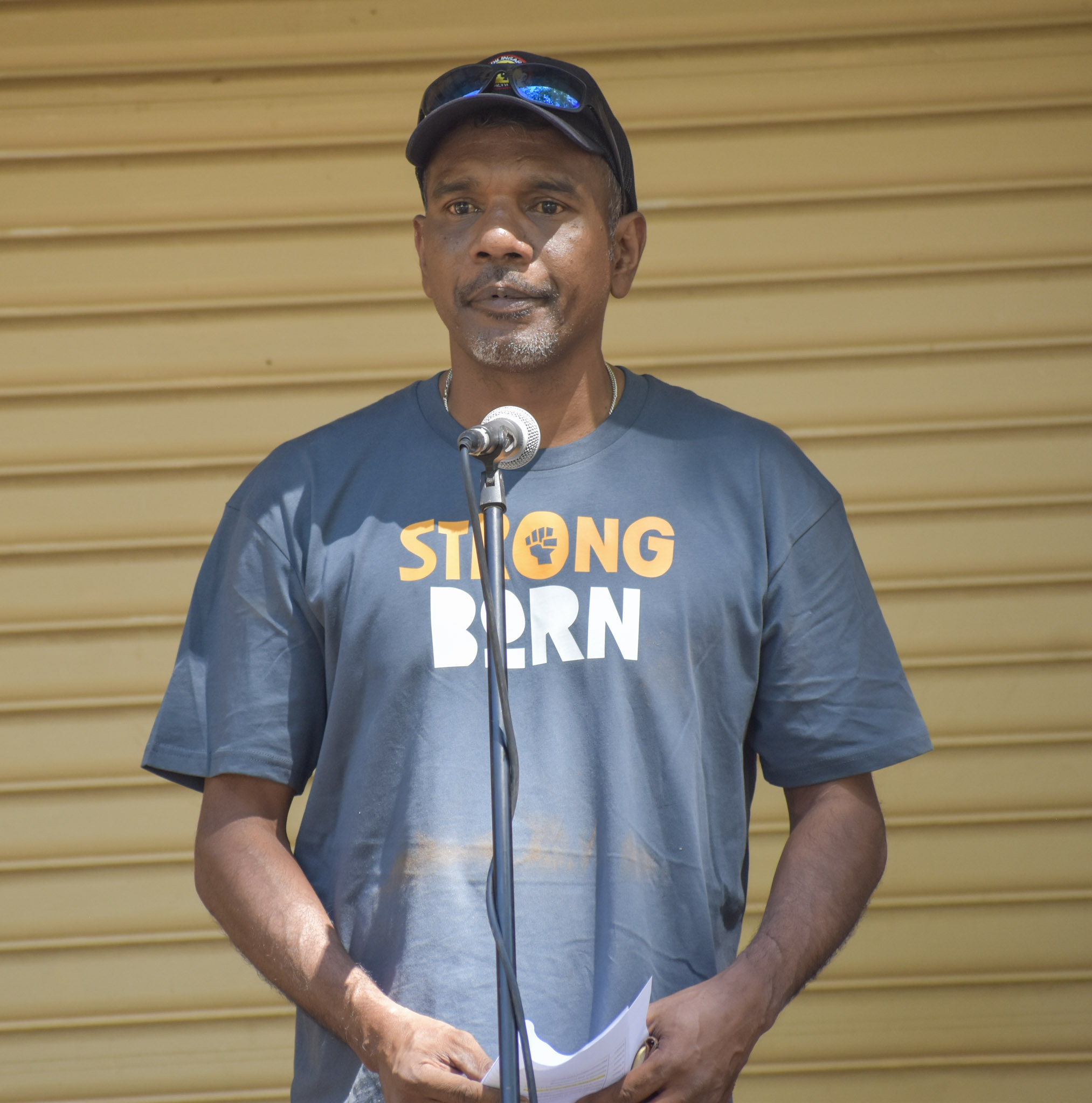 Aboriginal male leader (Ishmael Croft) wearing a blue T-shirt and baseball cap stands behind a microphone.