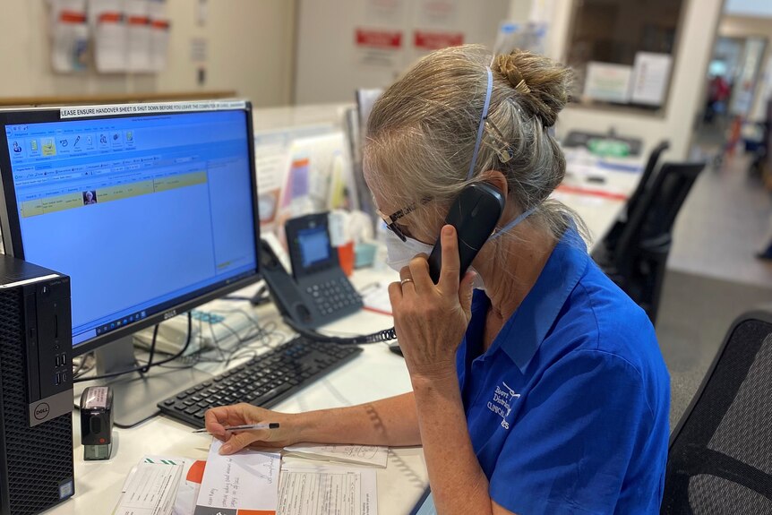 A woman with grey hair and a N95 mask sitting at a computer in an office, has a phone to her ear.