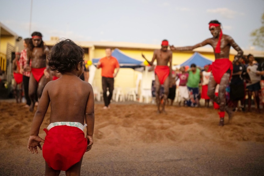 A toddler wearing a red cloth over their nappy watches Indigenous men wearing red dance in the background.