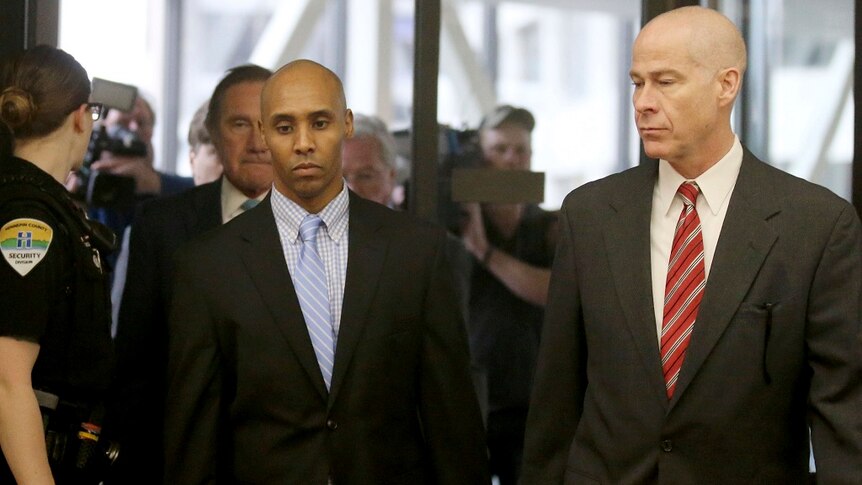 Two men in suits walk past a security guard on their way into a courtroom while being followed by a large group of media