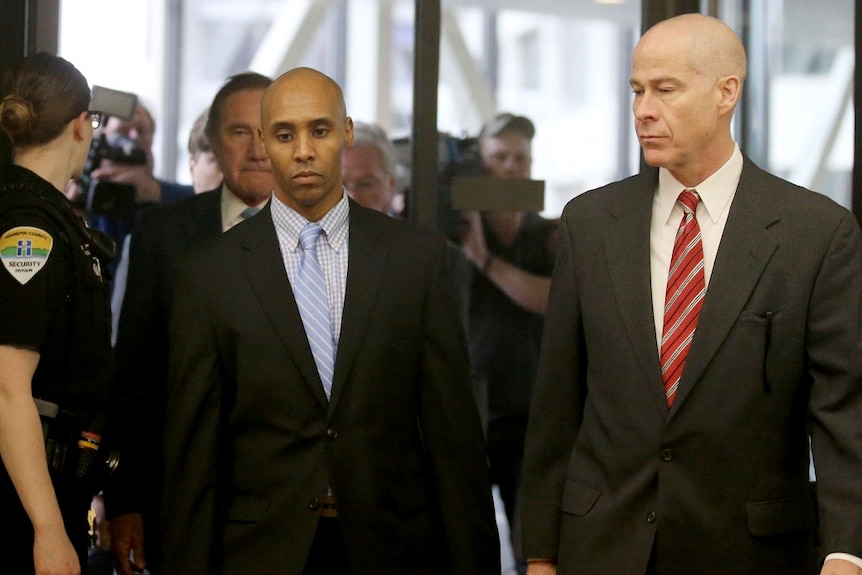 Two men in suits walk past a security guard on their way into a courtroom while being followed by a large group of media