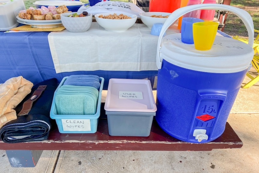 A picnic-table set up at the park, with supplied on the seat including a box of reusable clean wipes and an esky-urn of drinks.