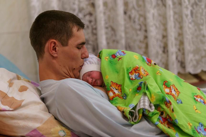A father lying on a hospital bed kisses the head of his newborn
