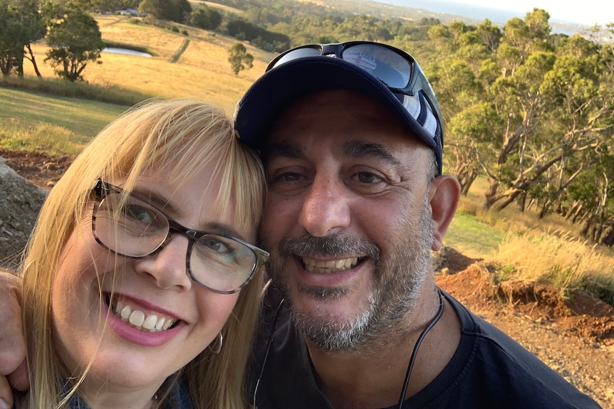 A blonde woman and man in a cap smile at a camera while taking a selfie.