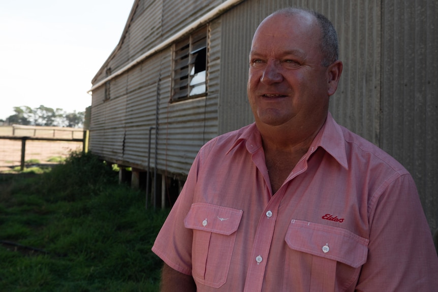 a man wearing a pink collared short with the logo 'elders' on the left breast pocket stand before an aging corrugated iron shed.